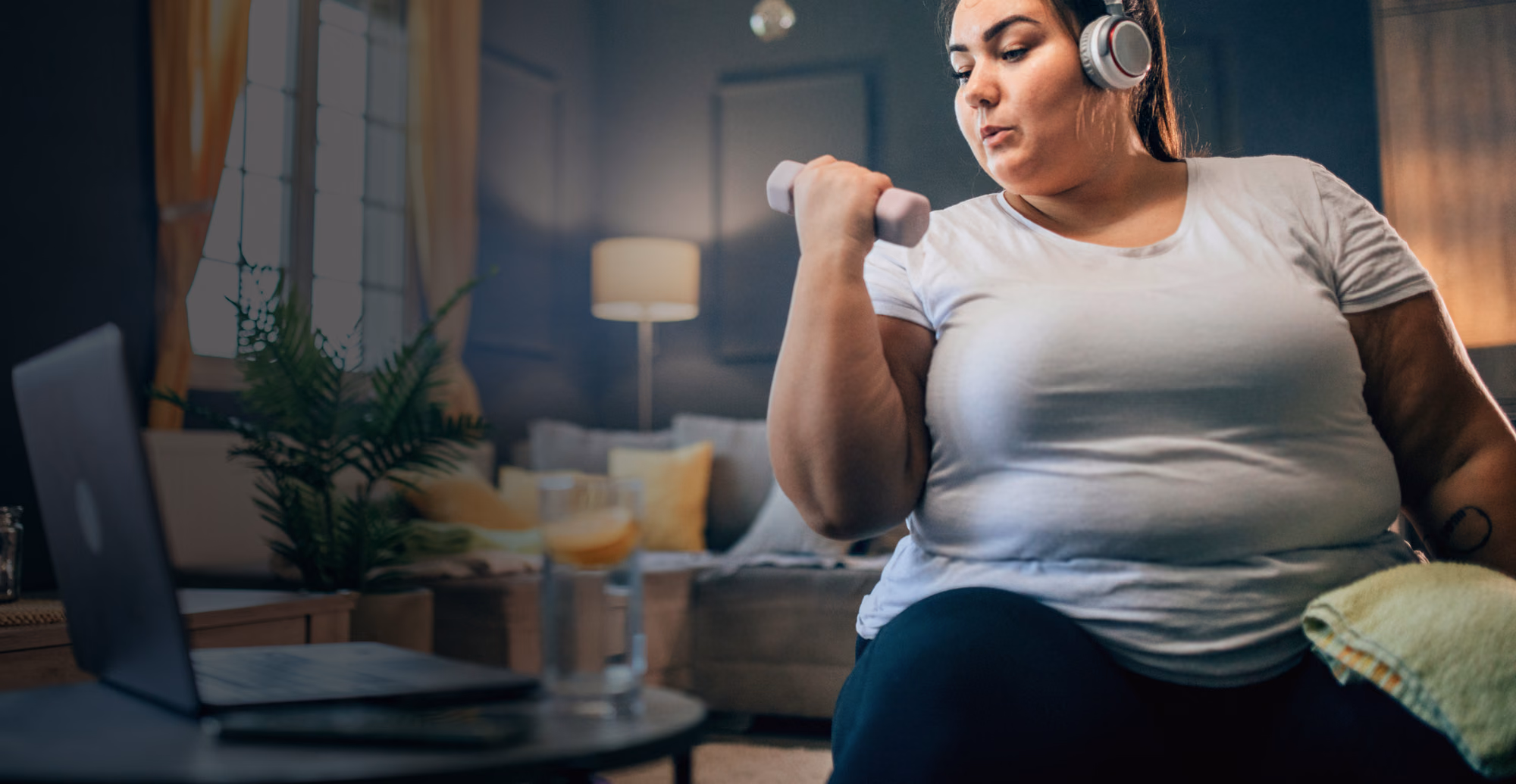 An obese woman in her early thirties doing bicep curls at home with water nearby. She’s sticking to the convenient activity plan she created with her virtual program care team.