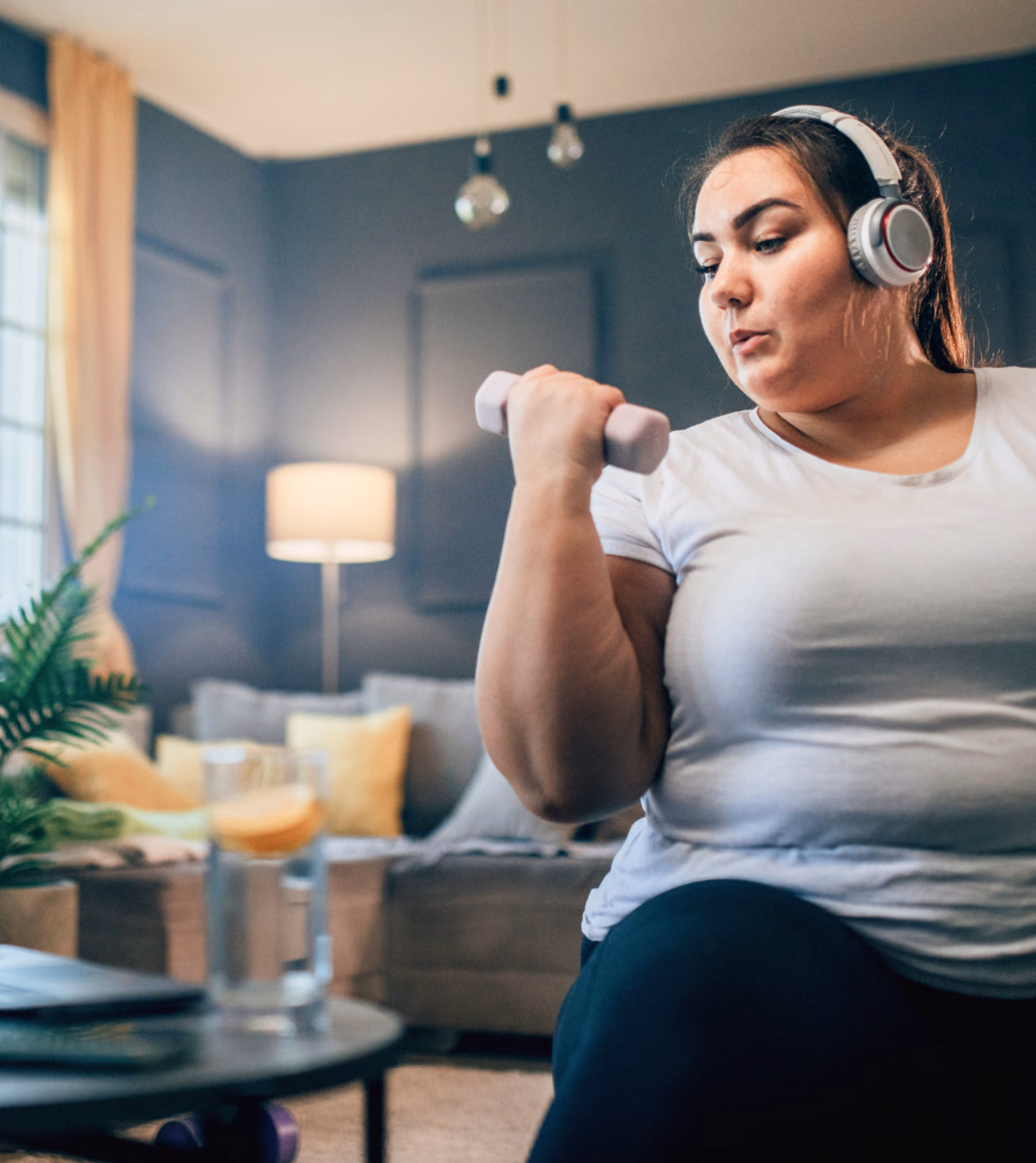 An obese woman in her early thirties doing bicep curls at home with water nearby. She’s sticking to the convenient activity plan she created with her virtual program care team.